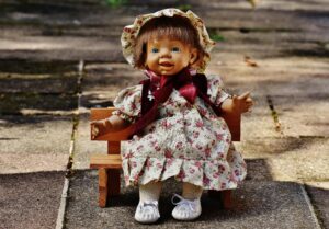 Cute smiling doll wearing a floral dress and bonnet, sitting on a wooden bench outdoors.
